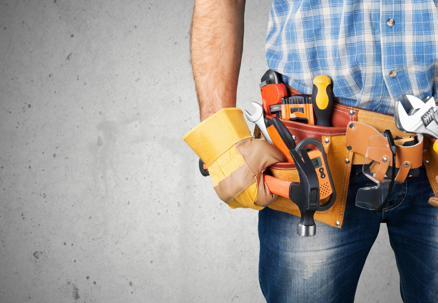 Person wearing a tool belt with various tools, dressed in jeans and a plaid shirt, standing against a gray wall.
